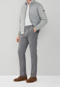 Giacca imbottita trapuntata grigio chiaro con collo alla coreana, camicia bianca abbottonata, pantaloni grigi a motivo scozzese e scarpe in camoscio marrone.