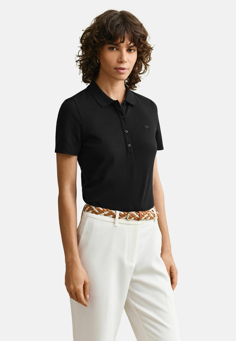 Femme aux cheveux bouclés vêtue d'un polo noir à manches courtes, pantalon blanc, et ceinture tressée beige et blanche, debout devant un fond uni.