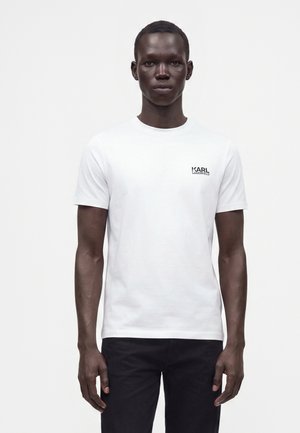 Uomo dalla pelle scura che indossa una t-shirt bianca Karl Lagerfeld e pantaloni neri, in piedi davanti a uno sfondo bianco semplice.