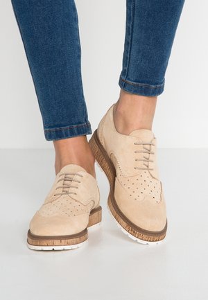Beżowe zamszowe buty brogue z perforowanymi detalami i drewnianą podeszwą noszone z podwiniętymi niebieskimi obcisłymi jeansami na stojącej osobie.