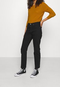 Jean noir coupe droite, associé à un pull en maille moutarde. La tenue est complétée par des baskets montantes noires à semelles blanches.