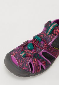 Lila och mönstrad sandal med justerbara remmar, en gummisula och en spärr för att säkerställa passformen; tillverkad av tvättbara material.