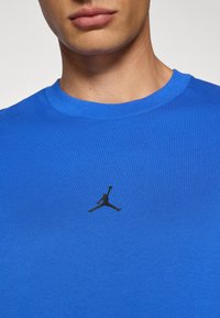 Tricou din bumbac albastru cu guler rotund și mâneci scurte. Logo-ul Jumpman de culoare neagră este centrat pe piept. Textură netedă.