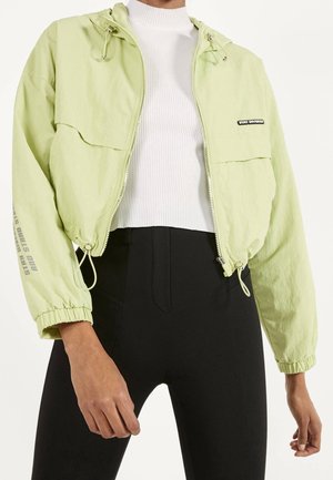 Person trägt eine hellgrüne, kurz geschnittene Jacke mit dem Schriftzug "STAY DIVERSE" und schwarze, hoch taillierte Hosen, kombiniert mit einem weißen, gerippten Top.