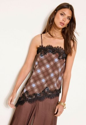 Femeie purtând o camisolă maro cu carouri și dantelă neagră la margină și pantaloni largi asortați, accesorizată cu brățări aurii și cercuri de urechi.