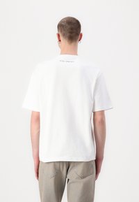 Camiseta de algodón blanca con mangas cortas, corte recto y detalle de texto en la parte superior de la espalda. La tela parece suave y lisa.