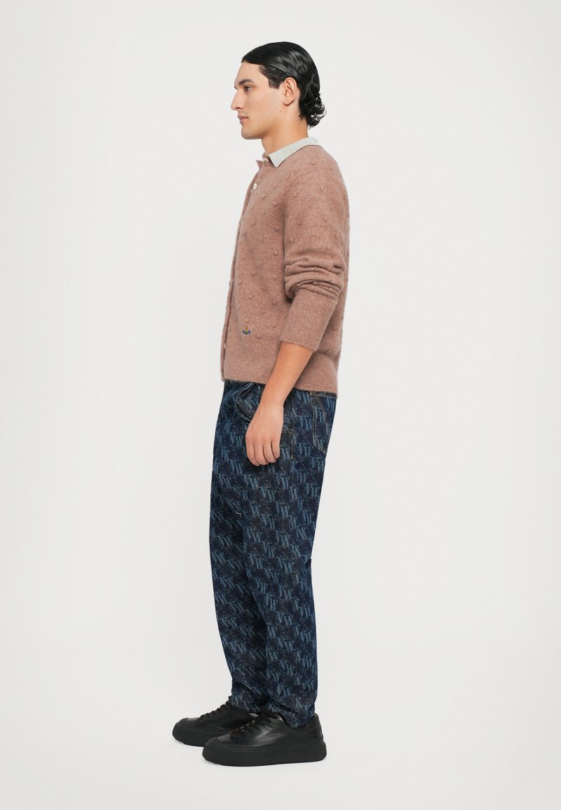 Bruine fuzzy cardigan met een gestructureerde afwerking, gecombineerd met marineblauwe patroonbroek en zwarte sneakers. Zijaanzicht, minimale achtergrond.