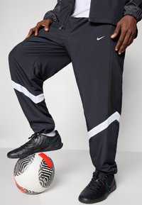 Sort Nike tracksuit bukser med hvide stribedetaljer og mesh-detaljering. Personen har sorte atletiske sko på og står på en mønstret fodbold.