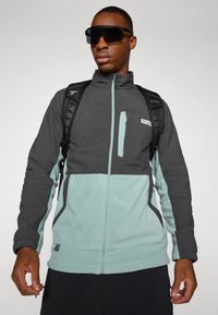 Chaqueta de forro polar gris y verde azulado con cremallera en la parte delantera, bolsillo en el pecho y puños elásticos. Llevada con gafas de sol oscuras y una mochila.