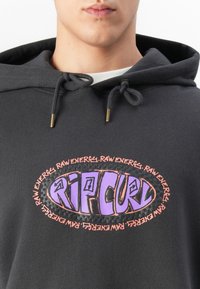Close-up van een persoon in een donkere hoodie met paarse "RIP CURL" tekst en "RAW ENERGY" in oranje rondom een ovale afbeelding op de borst.