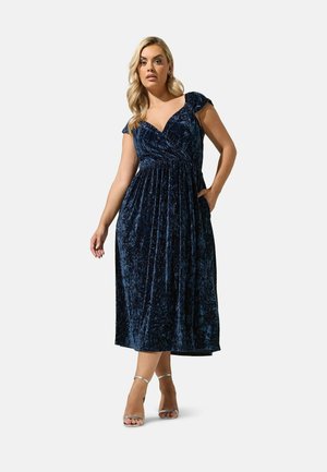 Mujer con cabello rubio que lleva un vestido midi de terciopelo azul marino con mangas tipo capa y sandalias plateadas de tacón alto, posando con una mano en el bolsillo.