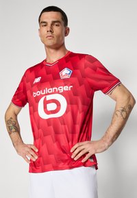 Rød herre fodboldtrøje med et struktureret geometrisk mønster, der har et hvidt logo og branding. Korte ærmer, V-hals og kontrasterende trimdetaljer.