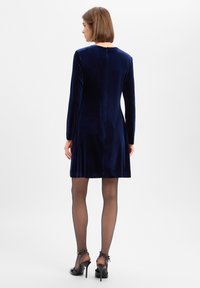 Marineblaues Samtkleid mit langen Ärmeln, Reißverschluss hinten, ausgestelltem Saum und glatter Textur. Kombiniert mit schwarzen transparenten Strumpfhosen und hochhackigen Schuhen.