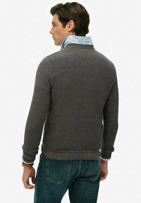 Szary sweter o fakturze z okrągłym dekoltem, noszony na jasnoniebieskiej koszuli w pasy, w zestawieniu z ciemnymi dżinsami. Widoczne detale przeszycia.