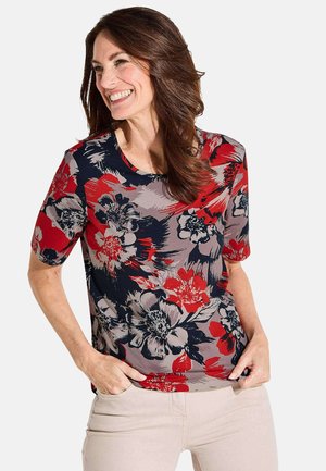 Haut à manches courtes floral avec une palette de couleurs bleu marine, rouge et gris, un col rond et une coupe décontractée, associé à un pantalon de couleur claire.