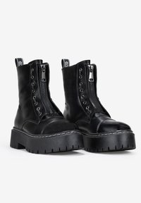 Botas de tobillo de cuero negro con suela gruesa, cremallera en la parte frontal y diseño con cordones. Presentan costuras blancas y tiradores para facilitar el uso.