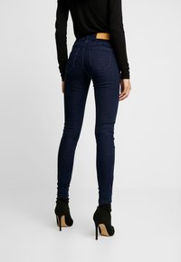 Person som bär mörkblå smala jeans och svarta high heeled ankle boots, stående mot en helt vit bakgrund.