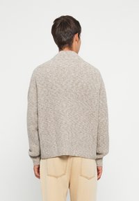 Mies, joka käyttää vaaleanbeigeä ribbikuvioista neulepaitaa ja vaaleita beigejä farkkuja, näkyy takaa pelkää valkoista taustaa vasten.