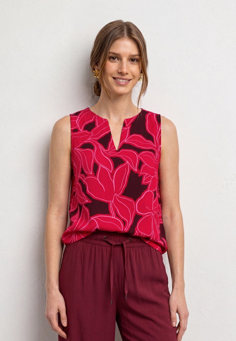 Femme portant un haut sans manches rouge et rose à motifs floraux avec un pantalon bordeaux à cordon, souriante et debout devant un mur blanc uni.
