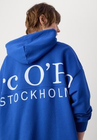 Blauer Hoodie mit großem weißem Grafiktext "c O'H STOCKHOLM" auf der Rückseite. Mit verstellbarem Kapuzenzug und lockerer Passform. Weicher Stoff.