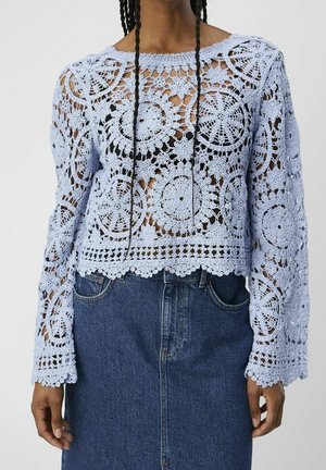 Top de encaje floral de crochet de manga larga en azul claro, usado sobre una camiseta negra, combinado con una falda vaquera de mezclilla azul oscuro de talle alto.