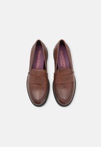 Mocassins en cuir marron avec une texture lisse, des bouts ronds et des coutures décoratives. L'intérieur présente une semelle intérieure violette avec le nom de la marque.