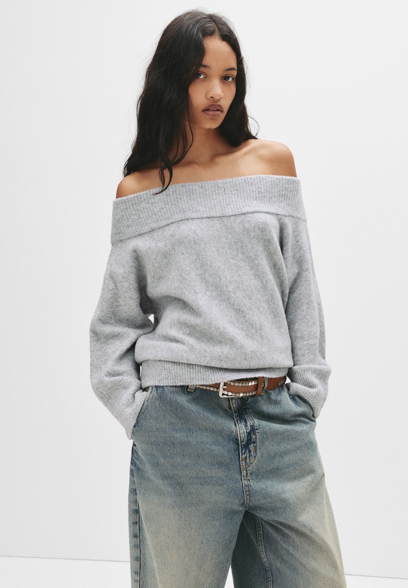 Grijze off-shoulder trui met ribbelneklijn, gecombineerd met lichtblauwe denim jeans met hoge taille en een bruine riem met decoratieve hardware.
