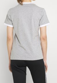 Šedé bavlněné tričko s krátkými bílými ribovanýma rukávy, kulatým výstřihem a volným střihem, v kombinaci s černými kalhotami se bílými bočními pruhy.