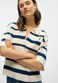 Beige stickad topp med marinblå ränder, kort ärm och krage. Har en V-hals och en liten broderad detalj på bröstet.