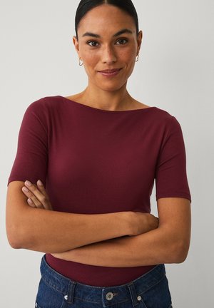 Kobieta z ciemnymi włosami w bordowej bluzce i niebieskich dżinsach stoi ze skrzyżowanymi ramionami, delikatnie się uśmiechając na jednolitym tle.