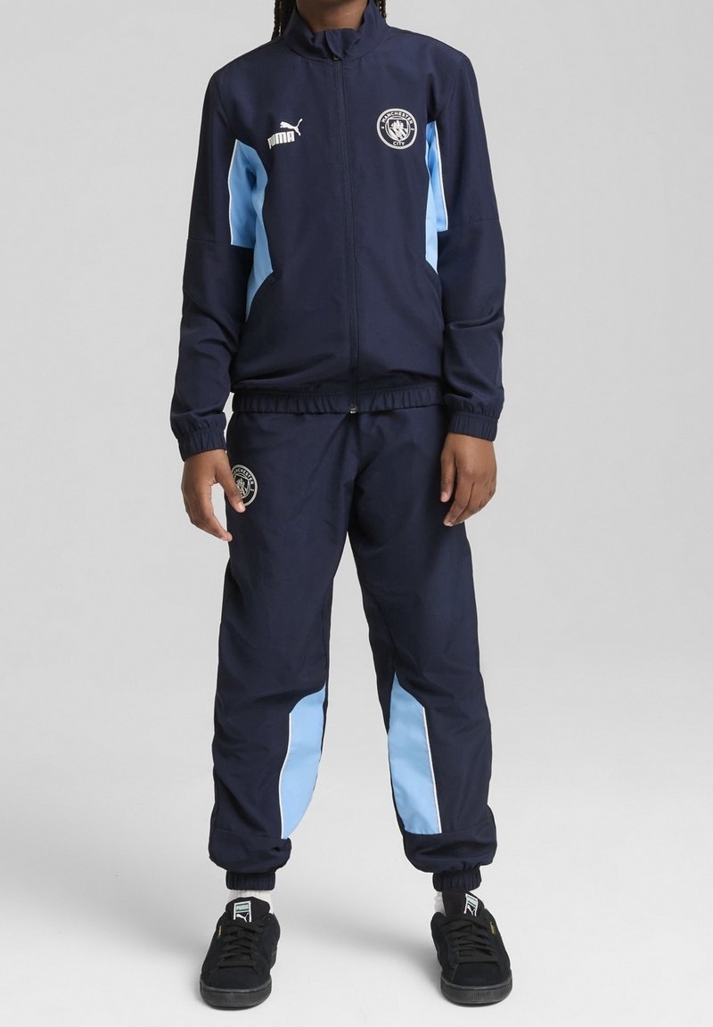 Survêtement marine avec des accents bleu clair, comprenant une veste zippée intégrale et un pantalon avec taille élastique. Comprend un logo sur la poitrine et la cuisse.