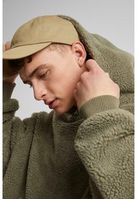 Olivengrüner Fleece-Hoodie mit hohem Kragen, kombiniert mit einer hellbraunen Baseballmütze. Verfügt über einen strukturierten Stoff und gerippte Bündchen.