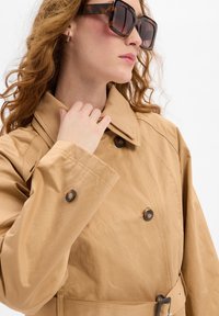 Beige dubbelzijdige trenchcoat met een grote kraag, bruine knopen en een riem. De stof heeft een gladde textuur. Bruin omhulde zonnebril.