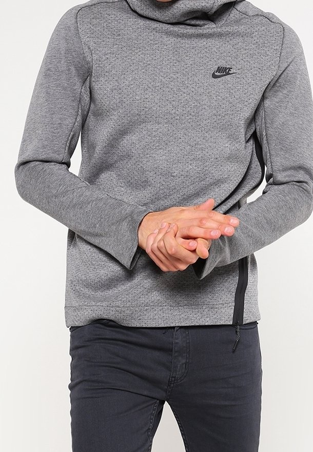Personne portant un sweat à capuche gris Nike à manches longues et un pantalon sombre, les mains jointes à la taille devant un fond uni.