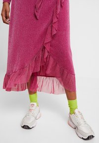 Vestido rosa brillante con dobladillo con volantes y superpuesto, combinado con zapatillas blancas y calcetines verde brillante. Primer plano de los detalles de la prenda inferior.