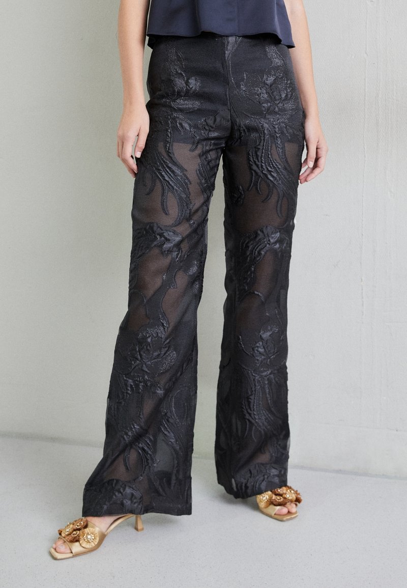 Mujer con pantalones anchos negros transparentes bordados y sandalias doradas de tacón con motivos florales, de pie contra una pared gris.