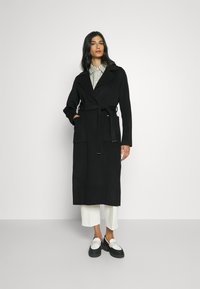 Abrigo de mezcla de lana negro con cintura ceñida por un cinturón, grandes solapas y dos bolsillos frontales. Combinado con una camisa a cuadros y mocasines blancos.