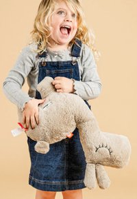 Zacht, beige knuffeldier in de vorm van een giraf met een ronde kop en ledematen, vastgehouden door een kind dat een grijs shirt en een spijkerbroek met bretels draagt.