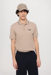 Beige polo triko s černým lemem, s logem na hrudi. Spárované s tmavě modrými kalhotami a odpovídající béžovou čepicí s plochým špičatým krempou.