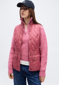 Γυναίκα με ναυτικό καπέλο, ροζ γιλέκο με καπιτονέ σχεδιασμό πάνω από ροζ πλεκτό πουλόβερ και μπλε τζιν, στέκεται μπροστά σε απλό φωτεινό φόντο.