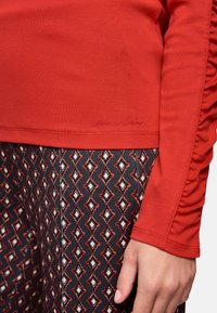 Red long-sleeve shirt with ruffled detail on the sleeve, paired with patterned black leggings featuring diamond shapes in cream and red.