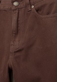 Jean en denim marron en gros plan montrant le bouton avant, la braguette zippée et la poche avant avec des détails de coutures visibles.