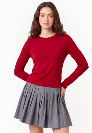 Donna con lunghi capelli castani che indossa una camicia rossa a maniche lunghe e una gonna grigia plissettata, in piedi con le mani sui fianchi.