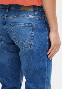 Jeans de mezclilla azul con bolsillos traseros con costuras en tono marrón contrastante y un acento en forma de pico. Textura suave y corte relajado.