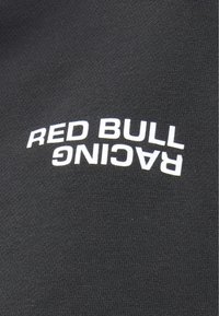 Μαύρο πουκάμισο από ύφασμα με το κείμενο «RED BULL RACING» σε έντονα λευκά γράμματα. Η υφή φαίνεται λεία και υφασμένη.