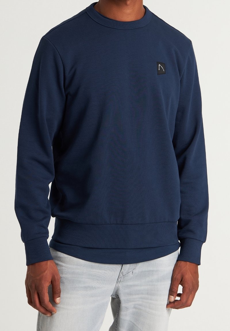 Homme portant un sweat-shirt bleu marine uni à manches longues avec un petit logo noir et blanc sur le côté gauche de la poitrine, associé à un jean gris clair.