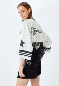Maglia bianca oversize con retro in pizzo nero, dotata di una grafica a stella e testo "Internazionale". Accenti a costine neri e maniche a righe.