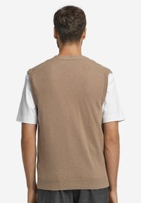 Brauner Strickwesten mit Rundhalsausschnitt und ärmellosem Design, über einem weißen T-Shirt getragen. Weiche Textur, lässige Passform, keine sichtbaren Muster.