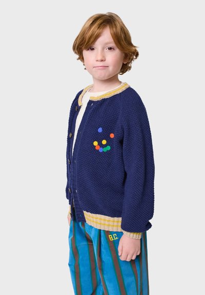 Mornarska pletena kardigan s žutim obrubom, koja ima šarene točkice na prsima, zatvaranje na gumbe i teksturiranu tkaninu. Uparen s prugastim hlačama.