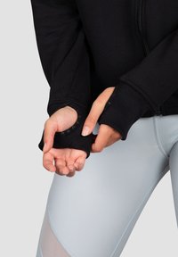 Melnā kapuces sporta jakā ar ribotām piedurknēm, kurās ir caurumi pirkstiem, kombinēta ar gaiši ziliem sporta legingiem. Gluda tekstūra un pieguļošs dizains.